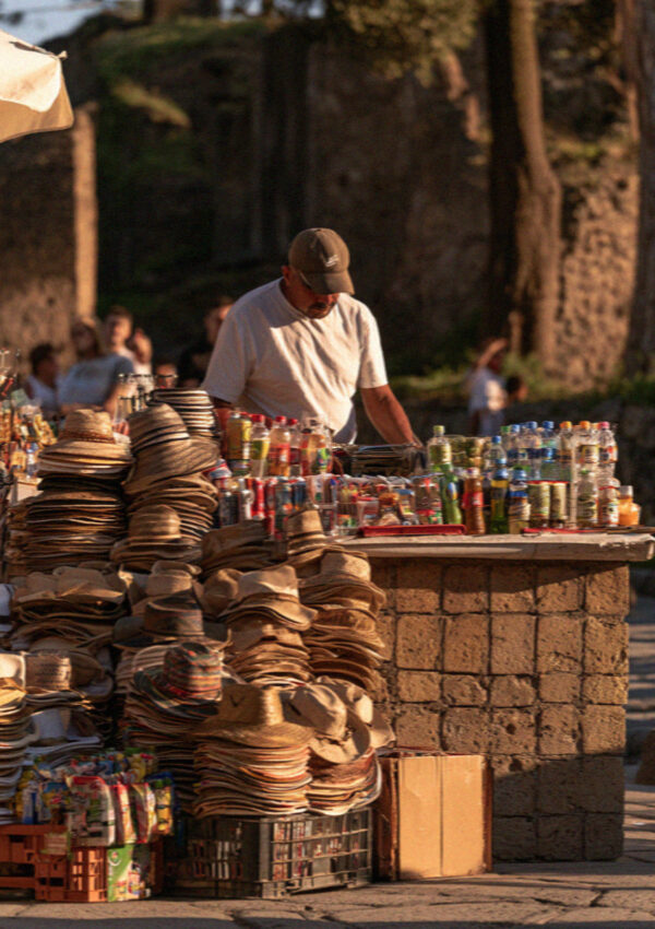 A souvenir seller with hats and cold drinks on a stall outside the site entrance in Pompeii in late afternoon light