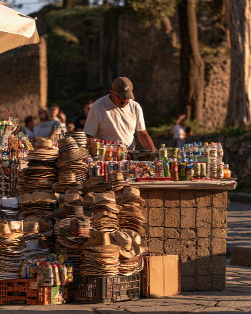 A souvenir seller with hats and cold drinks on a stall outside the site entrance in Pompeii in late afternoon light