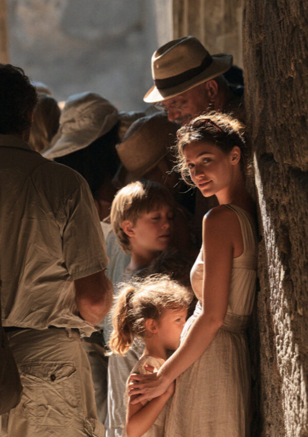 A group of tourists crowded into a narrow band of shade against a stone wall in Pompeii at midday