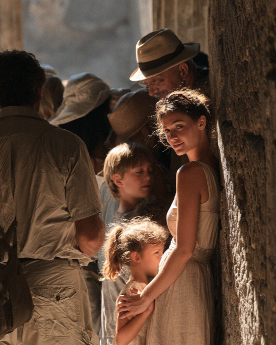 A group of tourists crowded into a narrow band of shade against a stone wall in Pompeii at midday