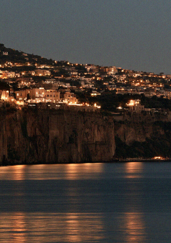 The lit town of Sorrento seen from the water at night, warm lights along the top of the dark cliff