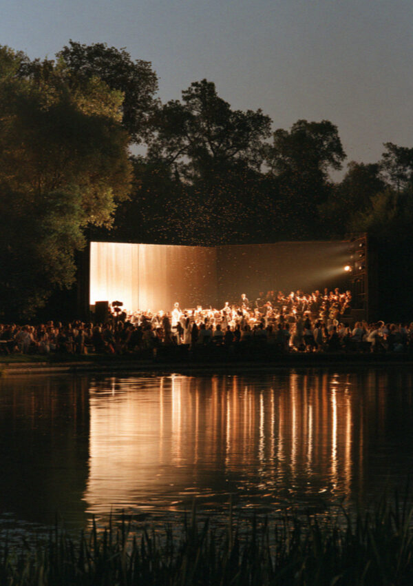 A lit open-air theatre on the shore of the lagoon at Torre del Lago in Tuscany at night, the audience in silhouette and the stage reflected in the still water