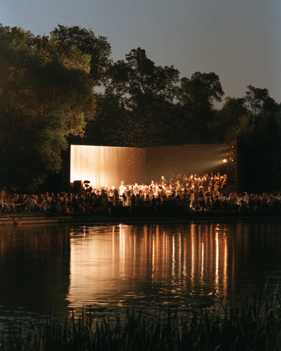 A lit open-air theatre on the shore of the lagoon at Torre del Lago in Tuscany at night, the audience in silhouette and the stage reflected in the still water