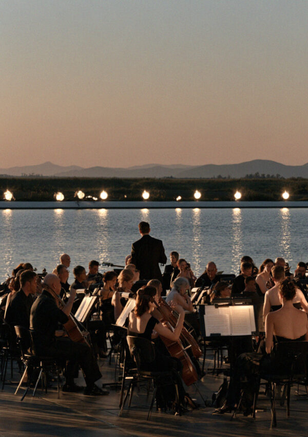 Musicians tuning in an open-air orchestra pit beside the lagoon at Torre del Lago in Tuscany at dusk, music-stand lights on and the audience seated behind
