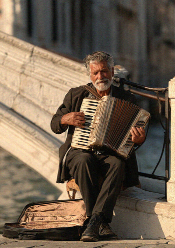 An older street accordionist playing beside a canal on stone steps in Venice