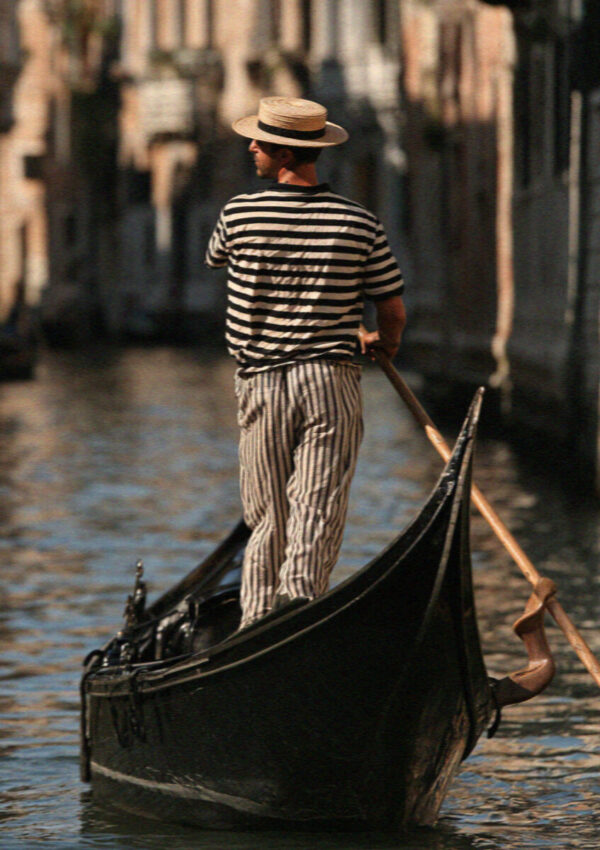A gondolier in a striped shirt and straw hat working the oar at the stern of his gondola on a canal