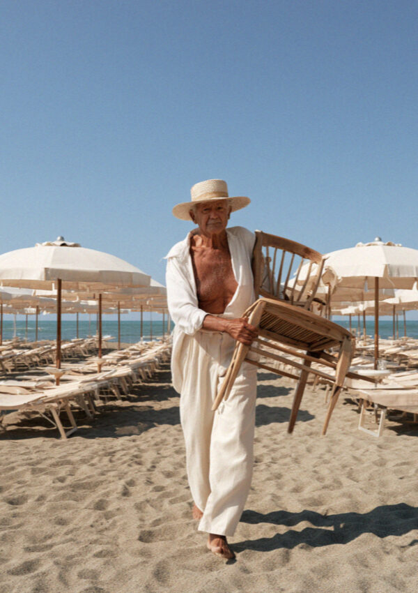 An older man in a straw hat carrying stacked wooden chairs across the sand between umbrella rows at a beach club on the Versilia coast in Tuscany