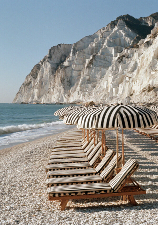 Ranked rows of striped umbrellas and wooden loungers on a Versilia beach in Tuscany below white quarried cliffs, no people