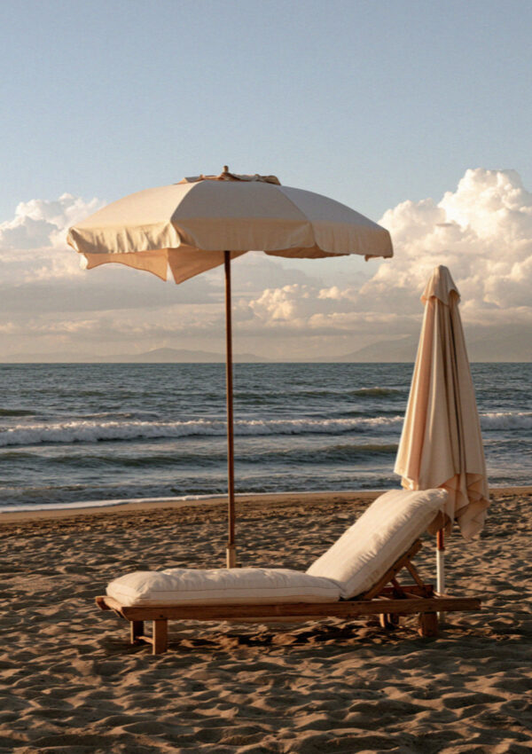A single wooden lounger and one open umbrella on empty sand on the Versilia coast in Tuscany in late afternoon light, the sea flat behind