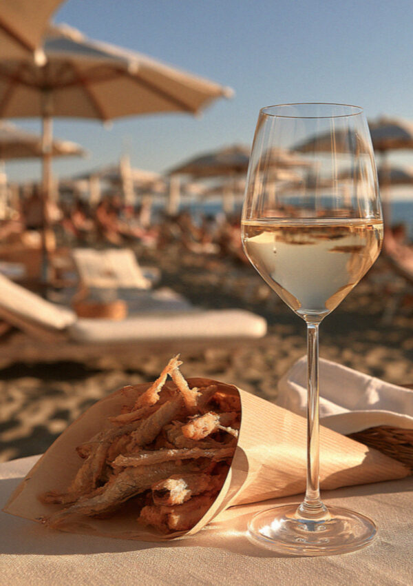 A paper cone of fried small fish and a cold glass of white wine on a beach club table on the Versilia coast in Tuscany, umbrellas soft behind