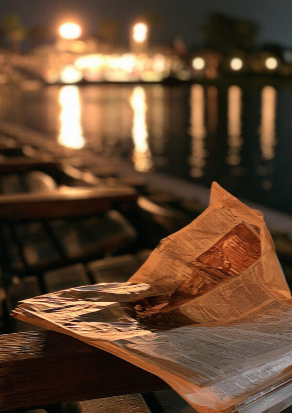 A paper cone of fried fish on newspaper at a table by the water at night on the Versilia coast in Tuscany, harbour lights soft behind