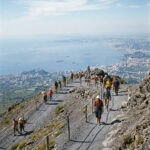 Hikers strung out along the zigzag grey gravel trail high on the bare upper slope of Vesuvius with the Bay of Naples hazy far below