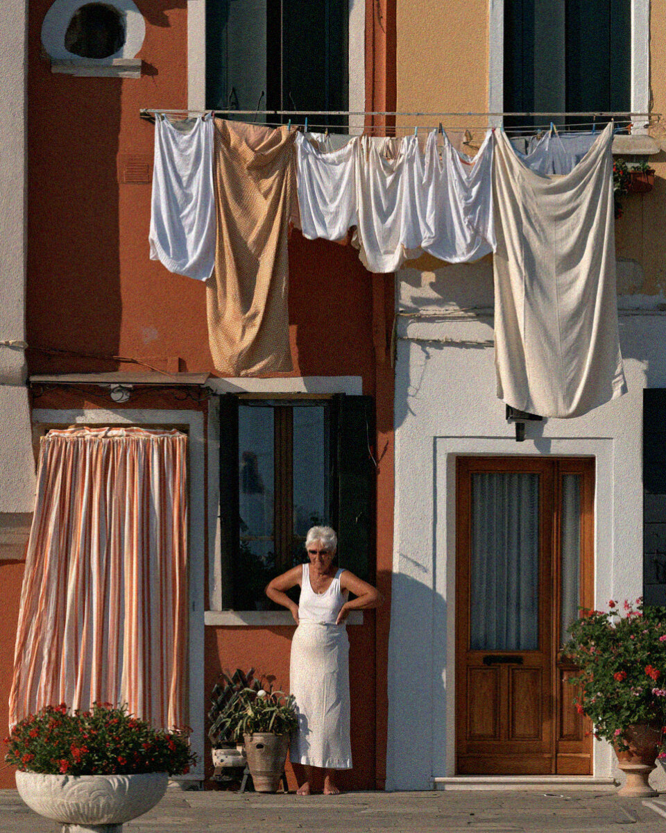 Victoria van der Berg in a peach dress seated beside a Burano canal with a moored boat.