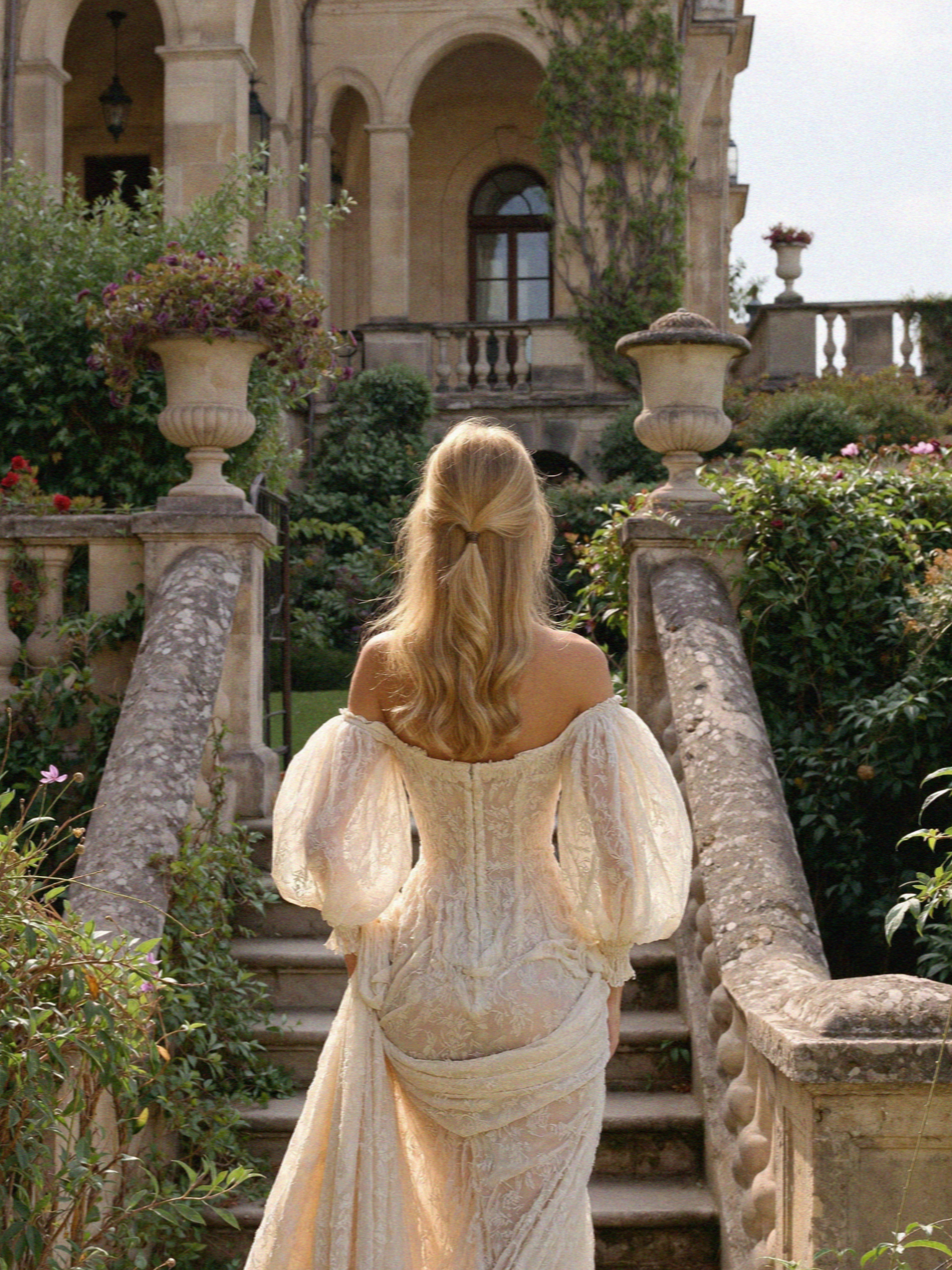 Victoria van der Berg in an off-shoulder lace wedding gown descending the stone garden steps of a villa on Lake Como