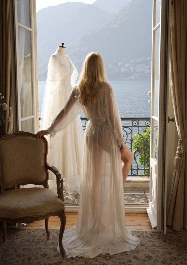 Victoria van der Berg in a silk robe getting ready by a hotel window on Lake Como with her gown hanging behind