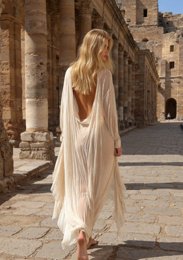 Victoria van der Berg in a long cream dress walking away down a colonnaded portico of weathered columns in Pompeii