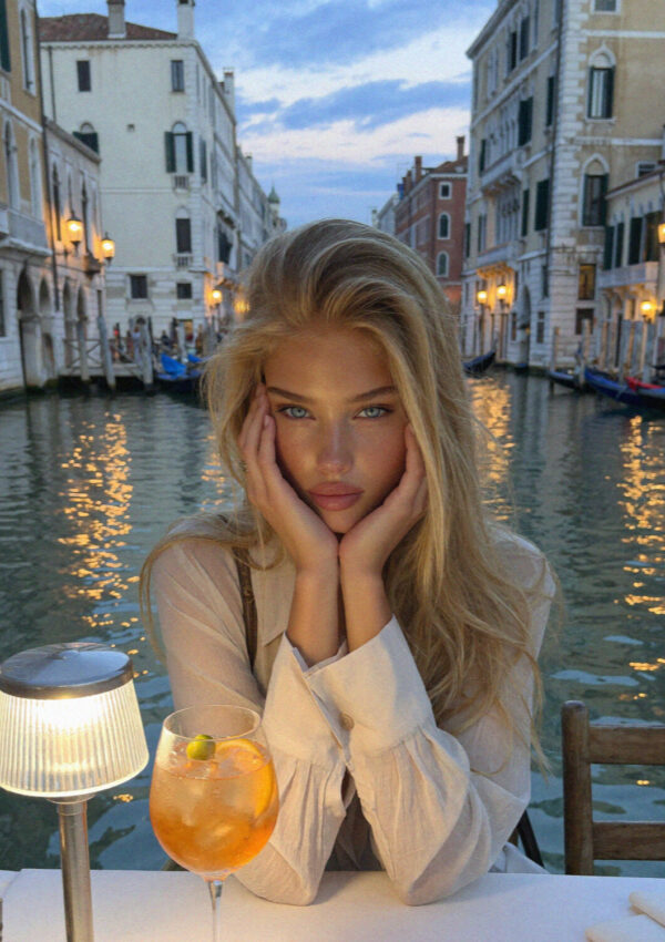 Blonde woman in cream resting her face in her hands at a Grand Canal table with a spritz at dusk, the Salute behind