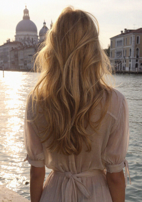 Blonde woman from behind in a cream dress looking over a Venice canal at golden hour