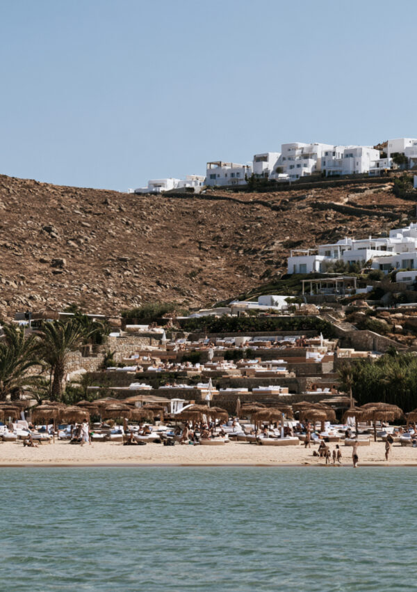 A Mykonos beach club of thatched umbrellas and loungers seen from the water below white hillside houses