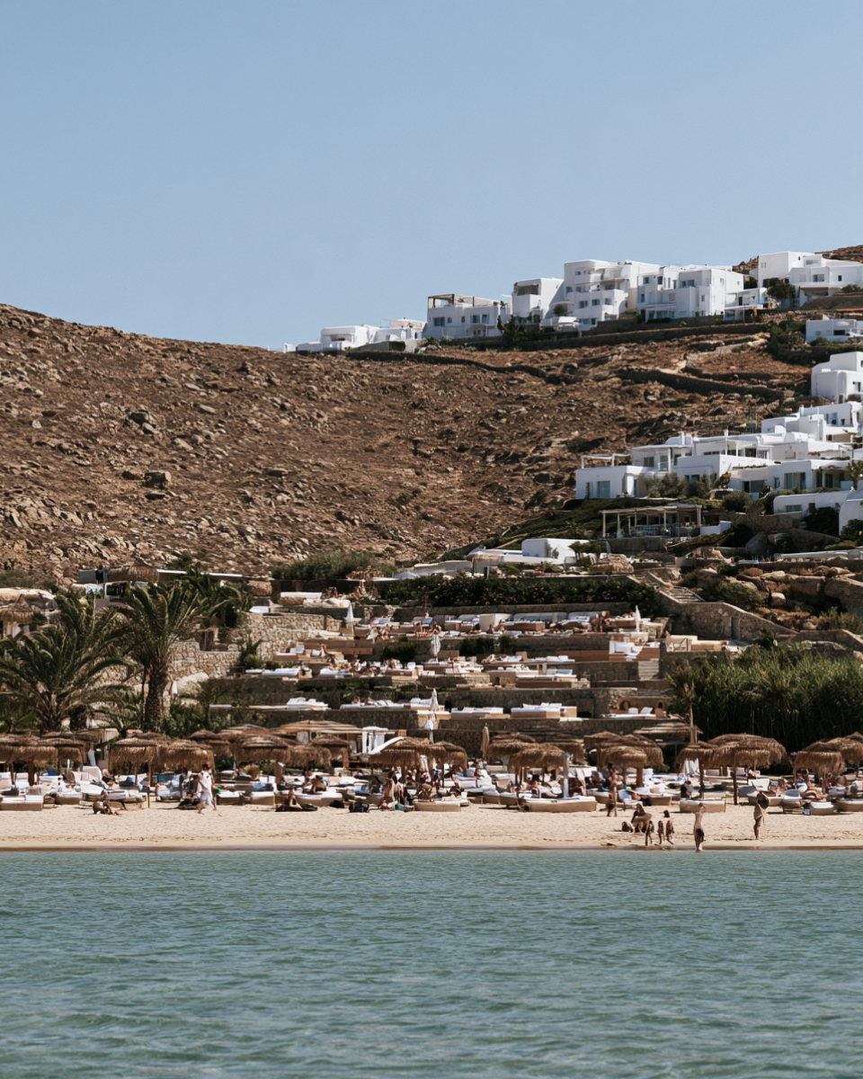A Mykonos beach club of thatched umbrellas and loungers seen from the water below white hillside houses