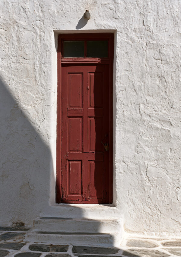 A dark red wooden door set into a thick whitewashed wall on Mykonos above stone paving
