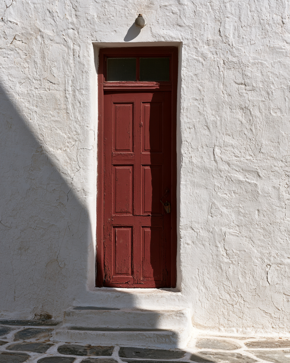 A dark red wooden door set into a thick whitewashed wall on Mykonos above stone paving