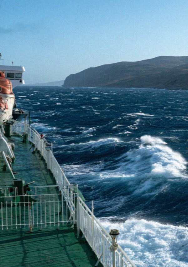 The deck railing and lifeboats of a ferry crossing a choppy Aegean sea towards Mykonos
