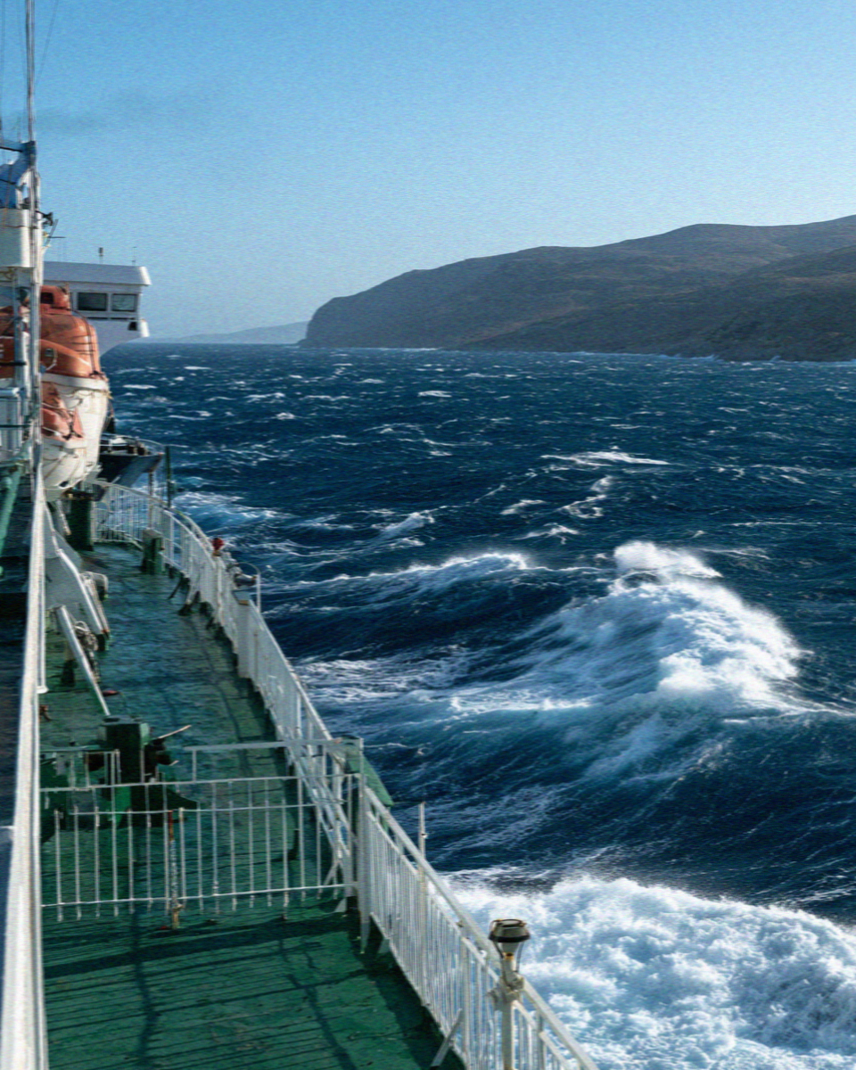 The deck railing and lifeboats of a ferry crossing a choppy Aegean sea towards Mykonos