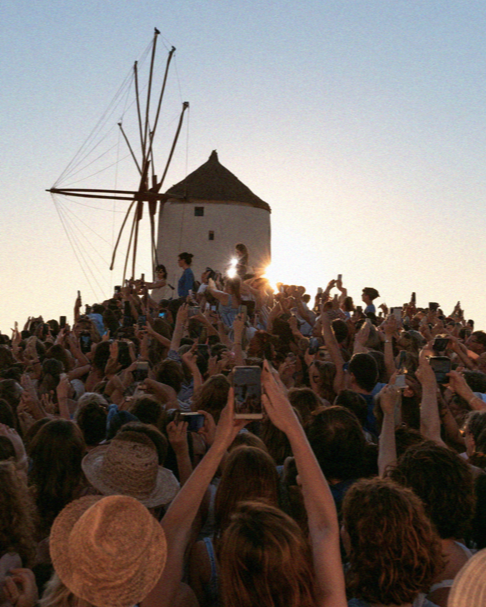 A crowd with raised phones photographing the windmills at sunset in Mykonos