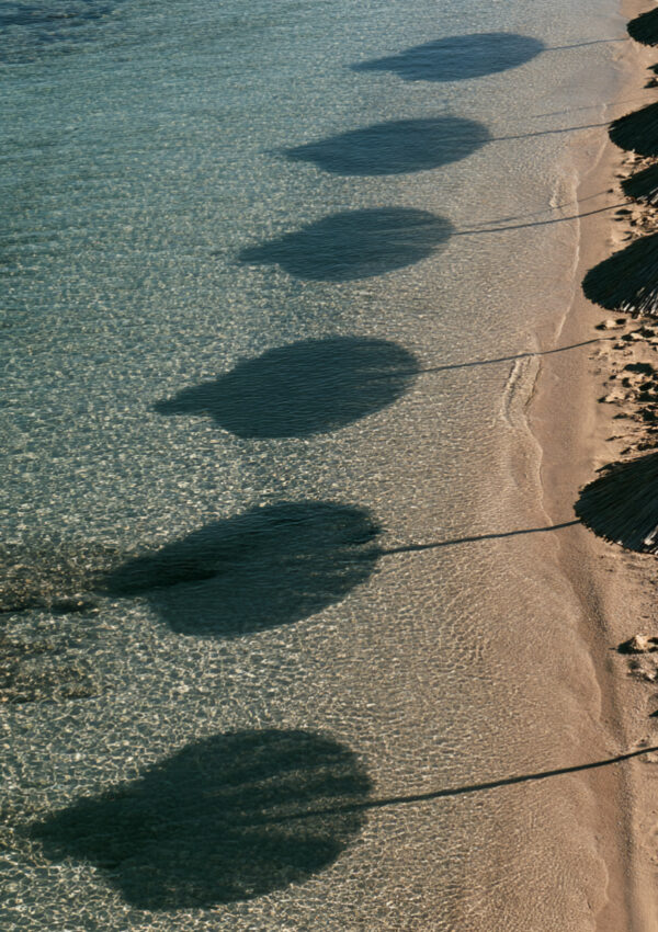A row of beach umbrella shadows falling across clear shallow water on a Mykonos beach