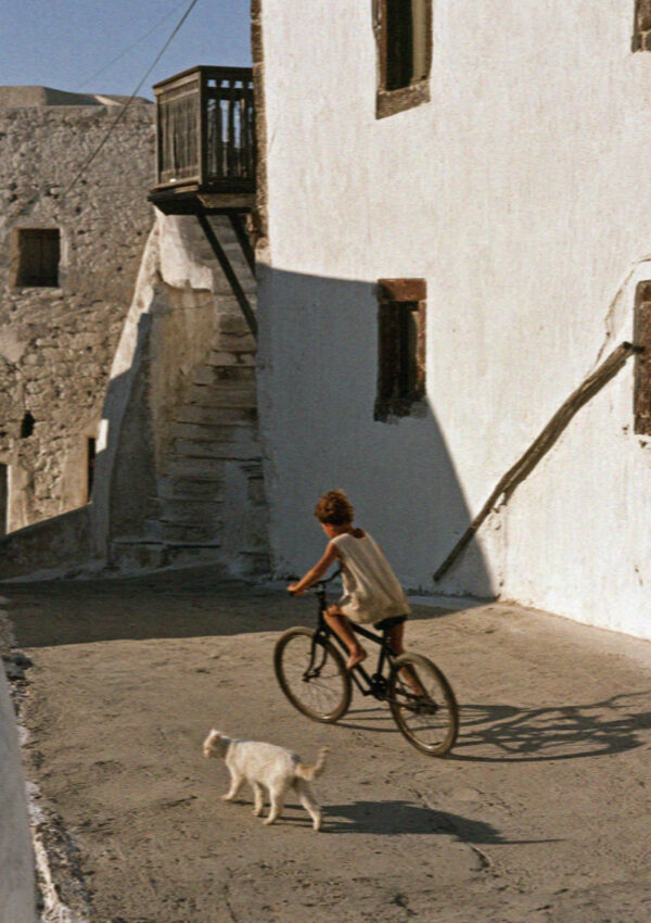 A boy riding a bicycle down a village lane with a dog running beside the front wheel in Santorini