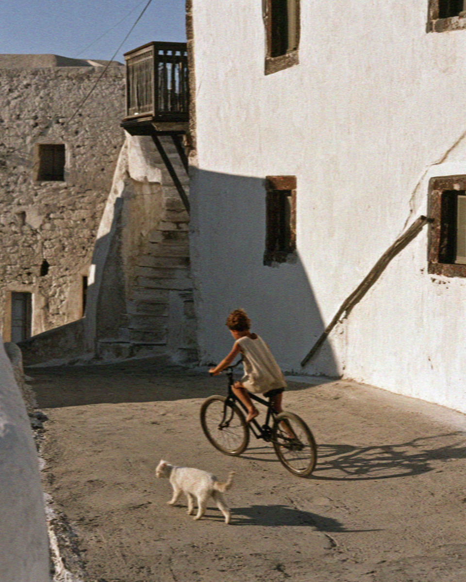 A boy riding a bicycle down a village lane with a dog running beside the front wheel in Santorini