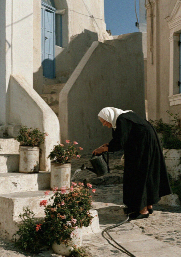 An older woman in black watering the plants on her front steps in a Santorini village