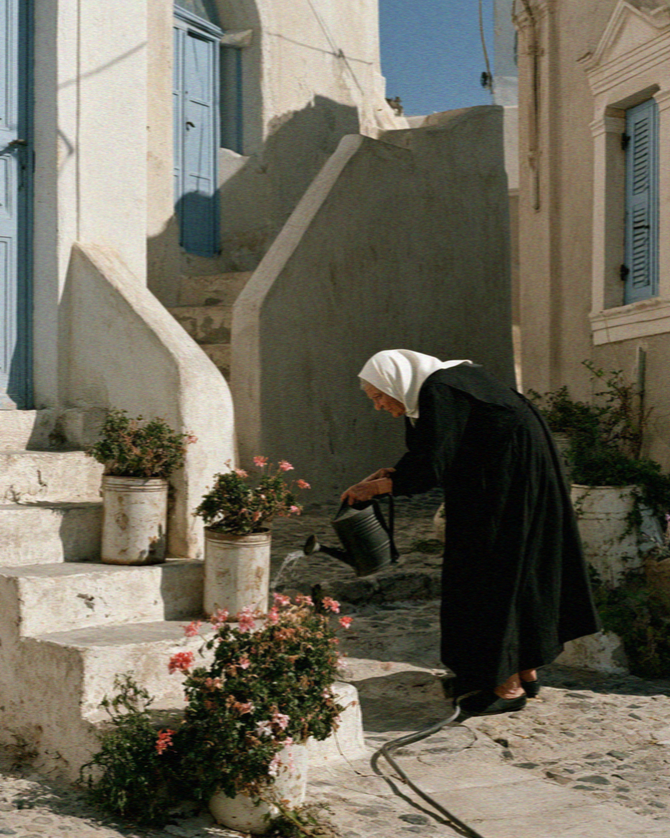 An older woman in black watering the plants on her front steps in a Santorini village