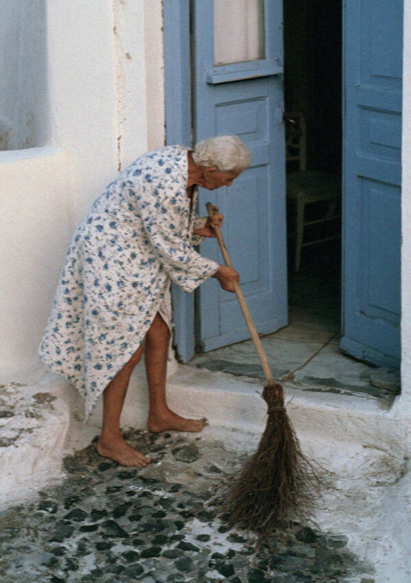 An older woman sweeping her doorstep with a twig broom at dawn in a Santorini village