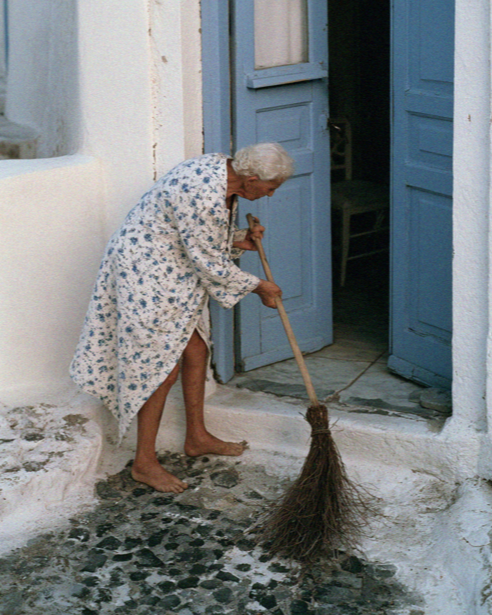 An older woman sweeping her doorstep with a twig broom at dawn in a Santorini village