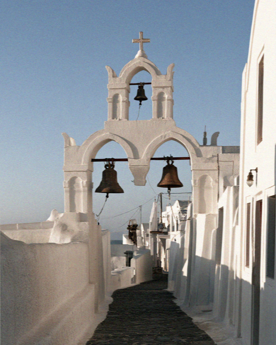 A whitewashed bell tower with bronze bells above a lane in Megalochori, Santorini