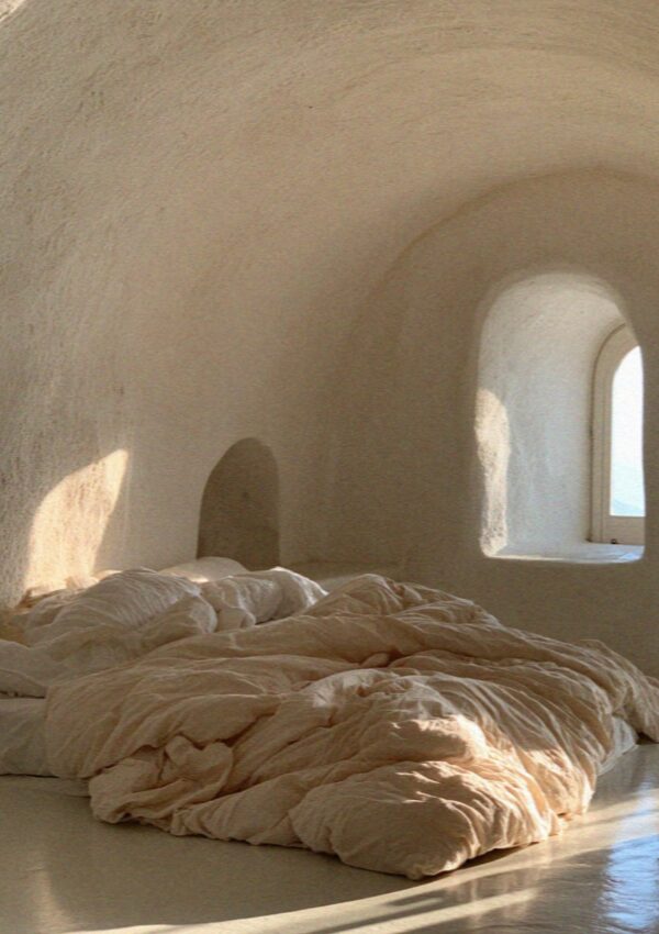 A whitewashed cave room carved into the caldera cliff on Santorini, with an unmade bed and an arched window at first light