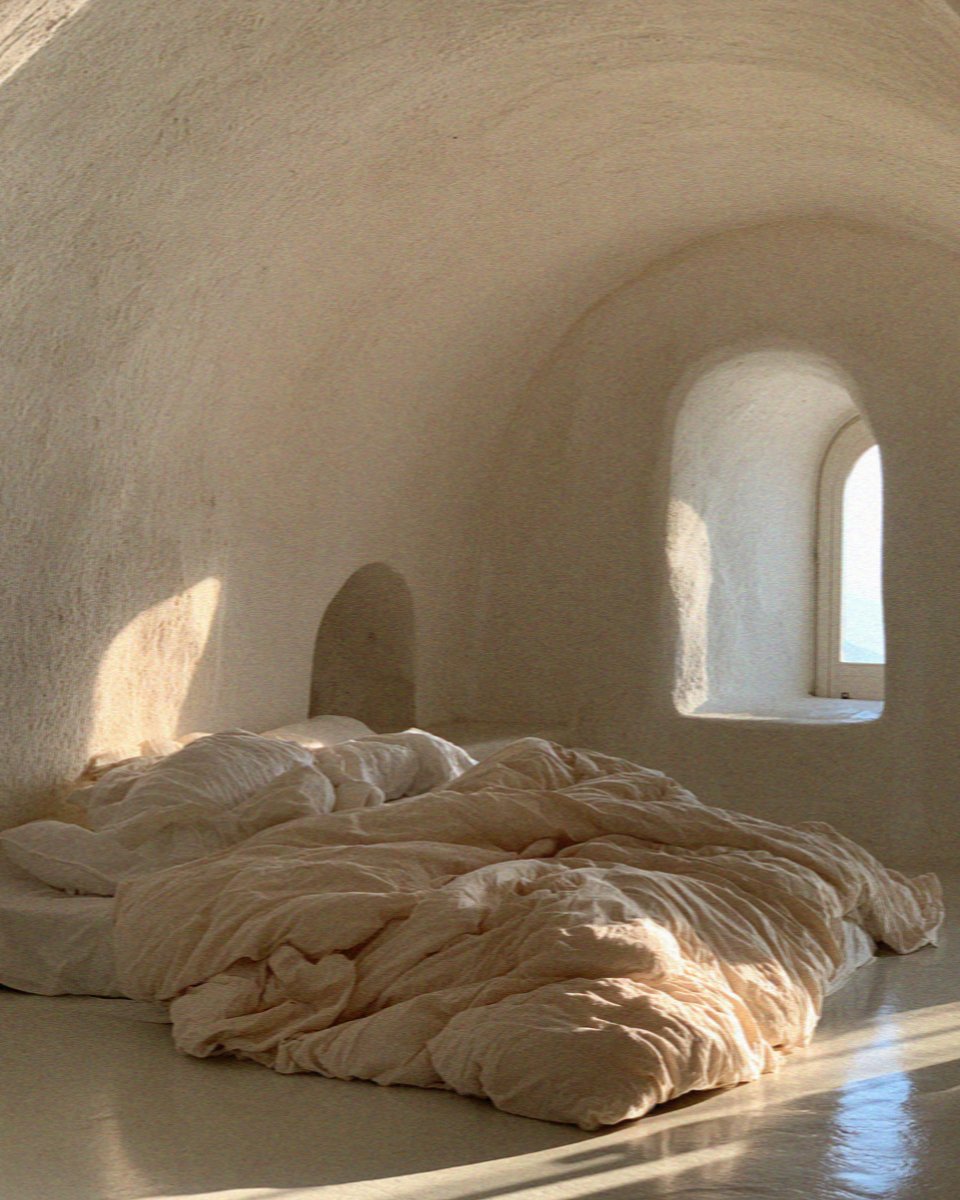 A whitewashed cave room carved into the caldera cliff on Santorini, with an unmade bed and an arched window at first light
