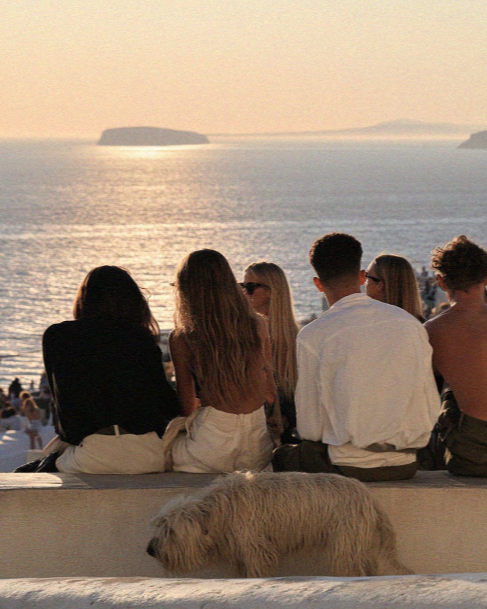 People and a dog sitting on a low wall at the top of Pyrgos watching the sunset over Santorini
