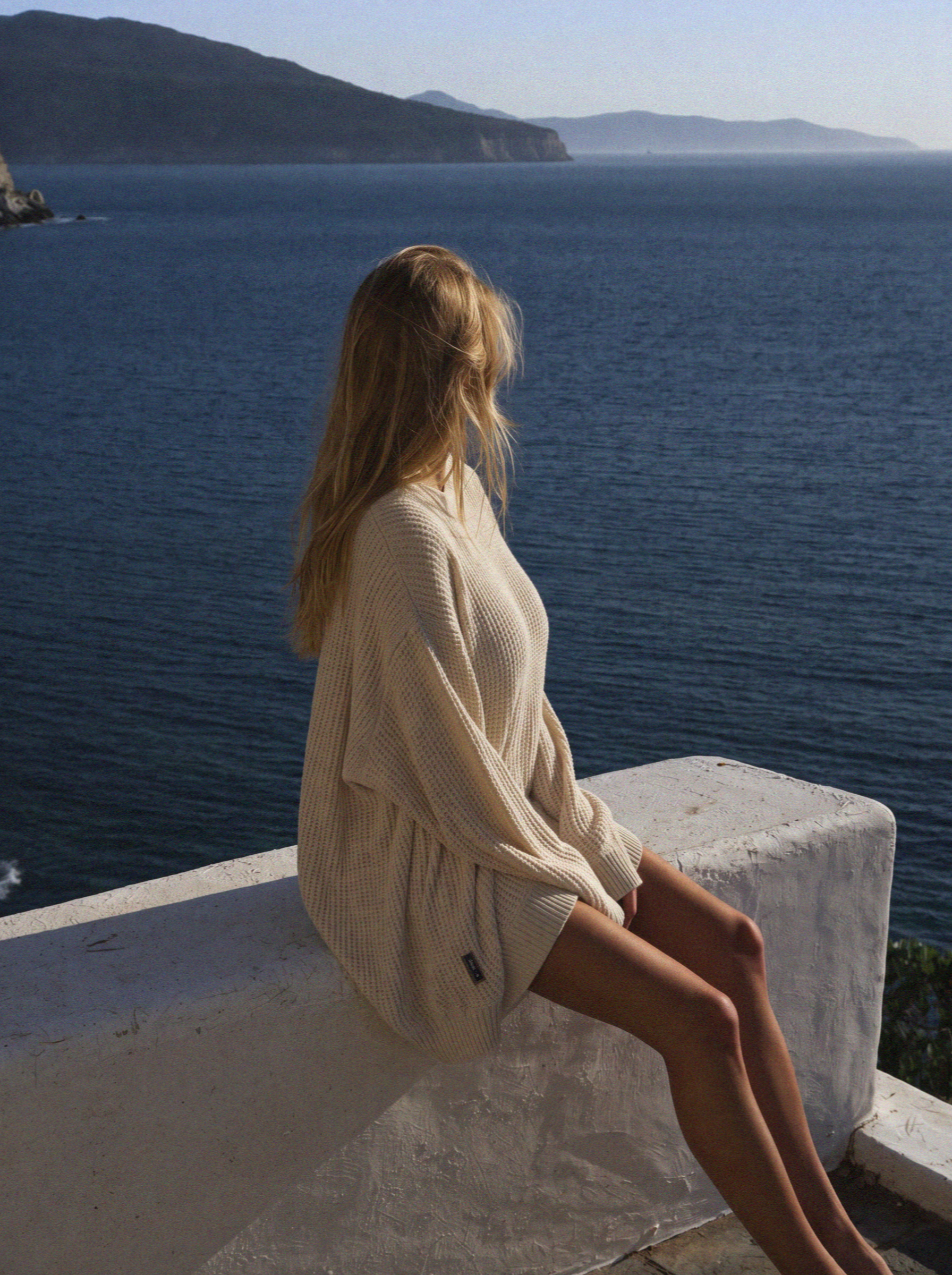 Victoria van der Berg in an oversized knit sitting on a villa wall above the sea at first light on Mykonos
