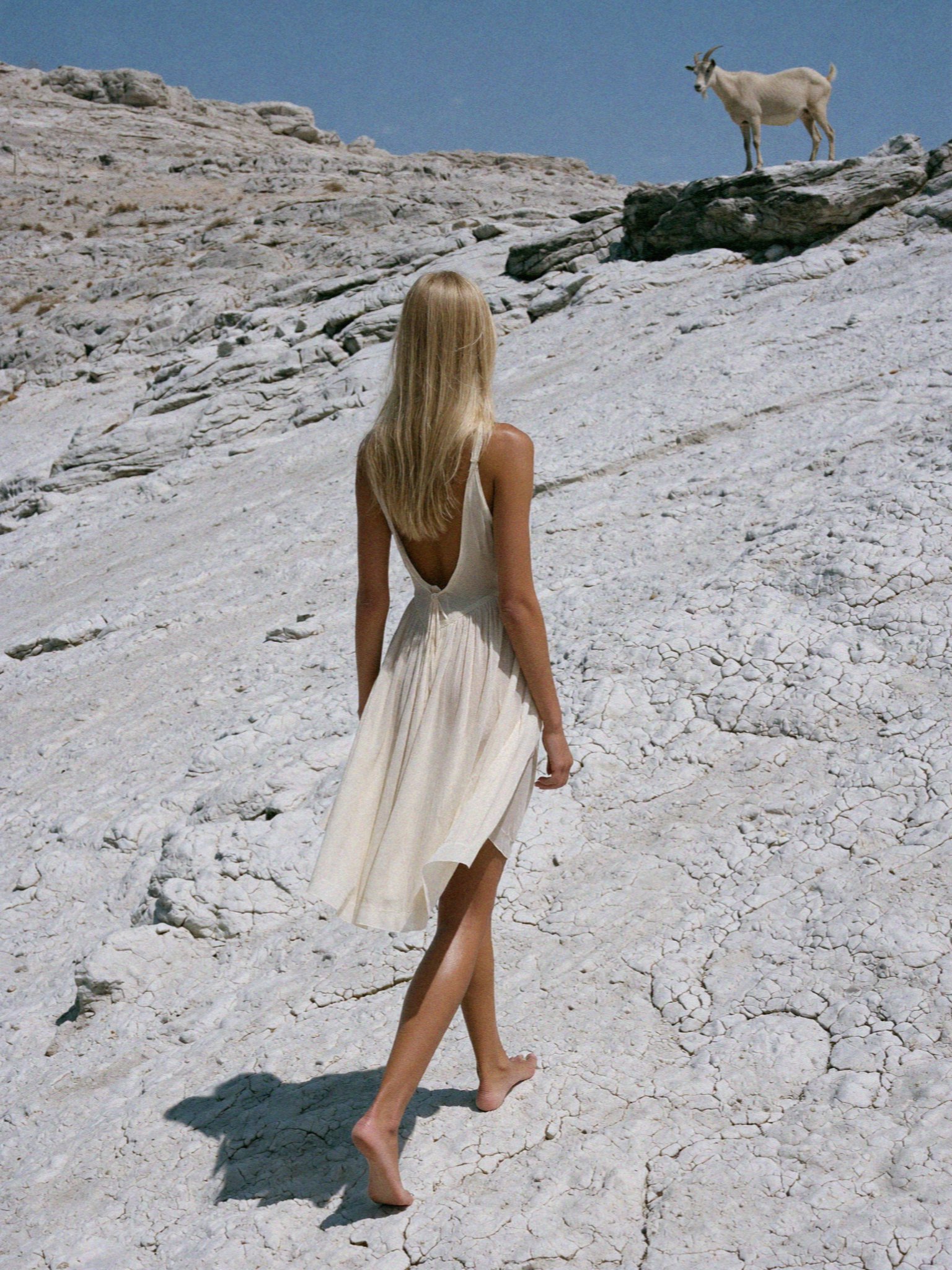 Victoria van der Berg in a white dress walking barefoot across white marble at the Marathi quarries on Paros, with a goat above
